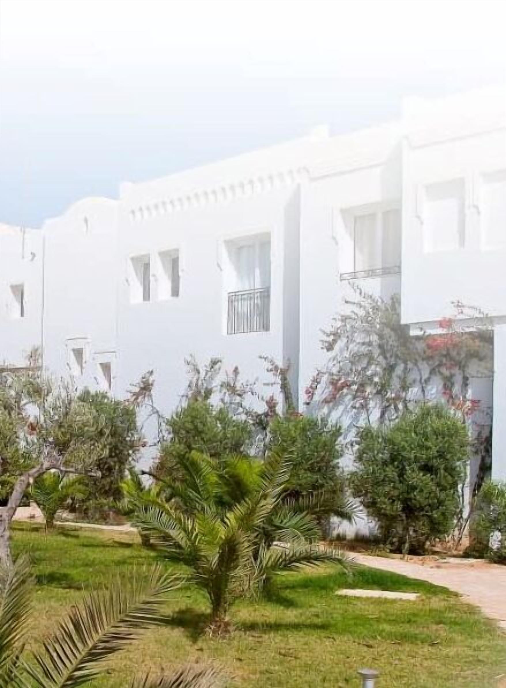 Un edificio blanco con múltiples ventanas y balcones, rodeado de vegetación y un camino.