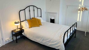 A neatly made bed with white bedding and two mustard-colored pillows, a bedside lamp, and a black metal headboard. - Cool 2 Bed House Hornchurch, Arcade Games, Parking (Hornchurch)