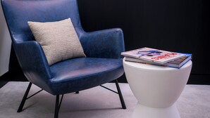 Un sillón de cuero azul con un cojín, una mesita blanca con revistas, y una gran obra de arte abstracta en la pared.