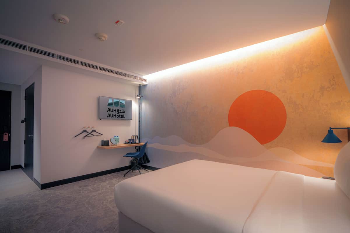 Una habitación de hotel moderna con una cama grande, un escritorio con una silla, un estante en la pared con artículos de aseo, y un mural grande en la pared.