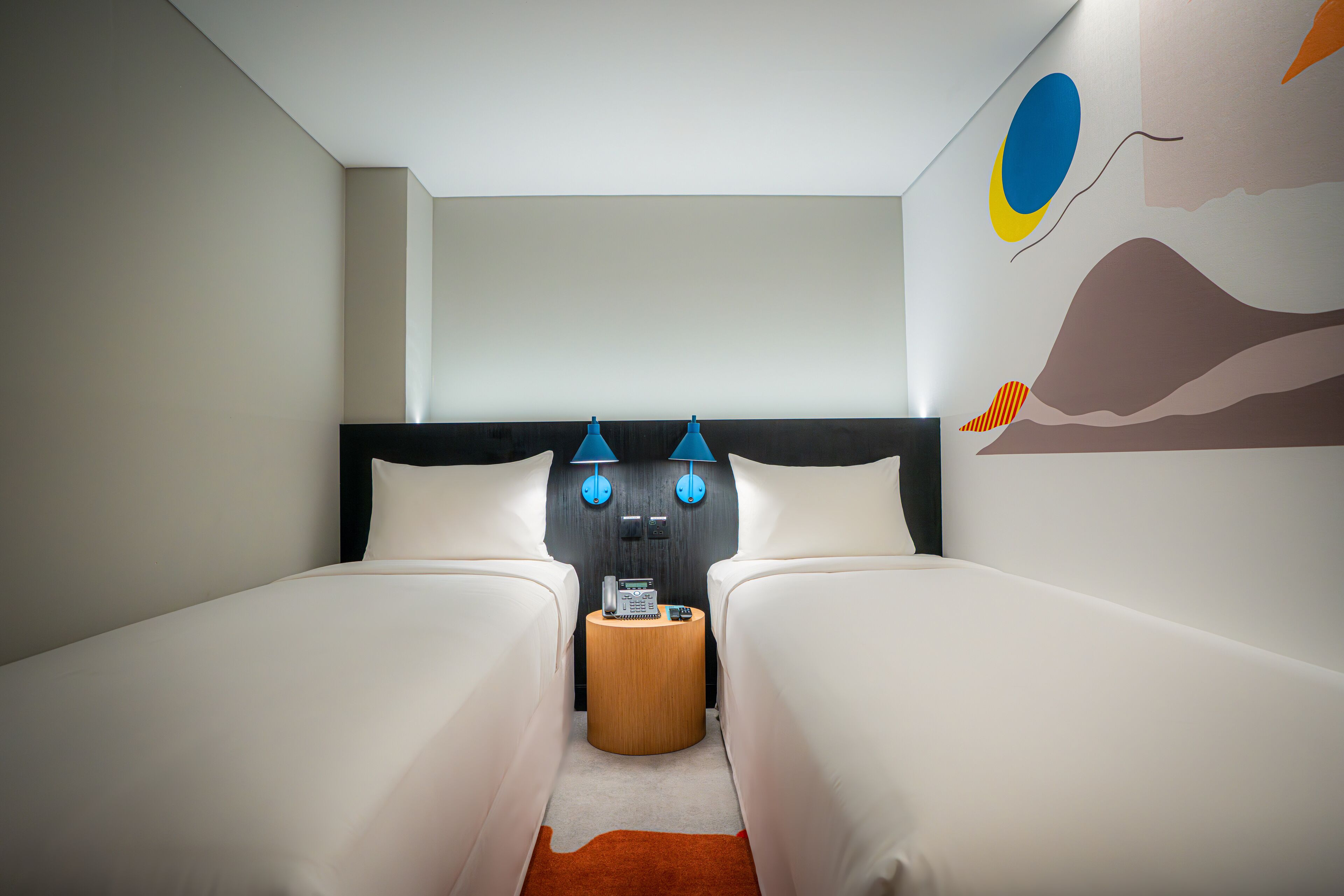 Habitación de hotel con dos camas individuales, una mesita de noche de madera, un teléfono y dos lámparas fijadas a la pared.