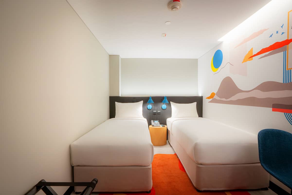 Habitación de hotel con dos camas individuales, una mesita, una silla y una pared con un mural colorido.