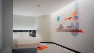A modern hotel room with a bed, a gray armchair, and a wall with colorful artwork.