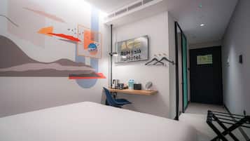 A modern hotel room with a bed, a desk with a chair, a wall-mounted shelf with toiletries, and a door leading to another room.