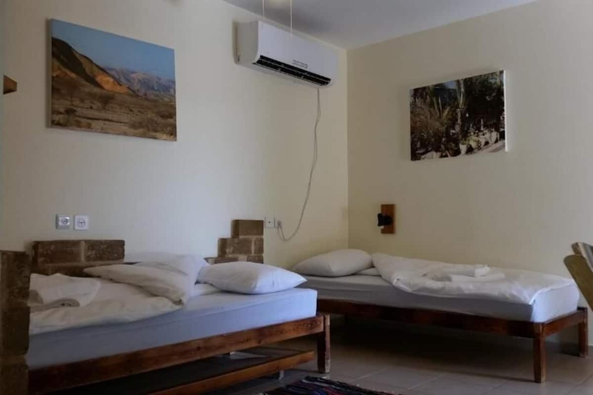 Ein Zimmer mit zwei Holzbett, einem kleinen Tisch und einem Stuhl. Zwei Wandklimaanlagen und eine Steckdose sind an der Wand angebracht.
