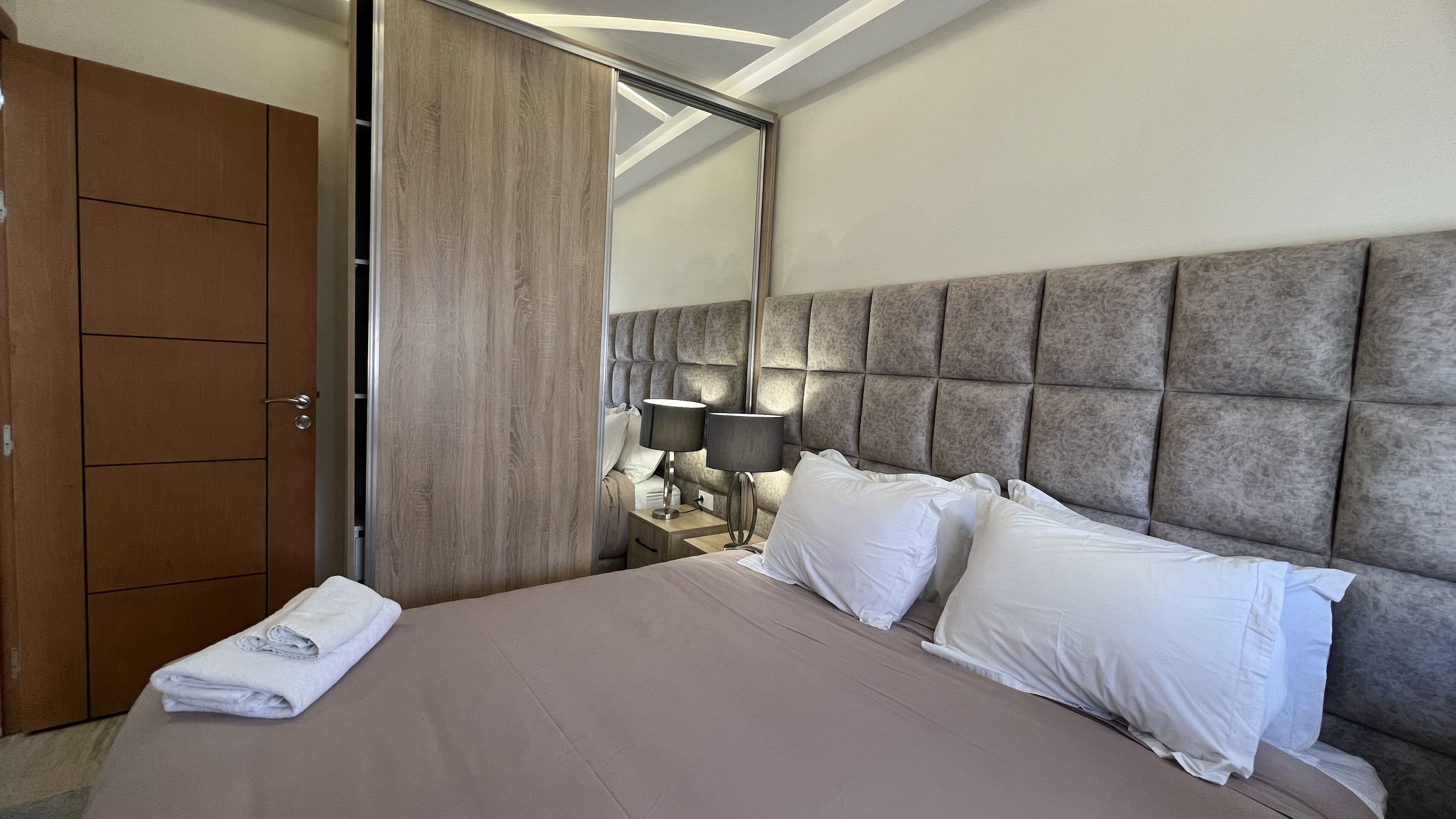 A hotel room with a large bed, a wardrobe, a nightstand with two lamps, and a mirror.