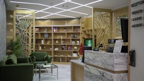 Un espace d’accueil doté d’un comptoir en marbre, de canapés verts et d’une bibliothèque en bois remplie de livres et d’objets décoratifs.