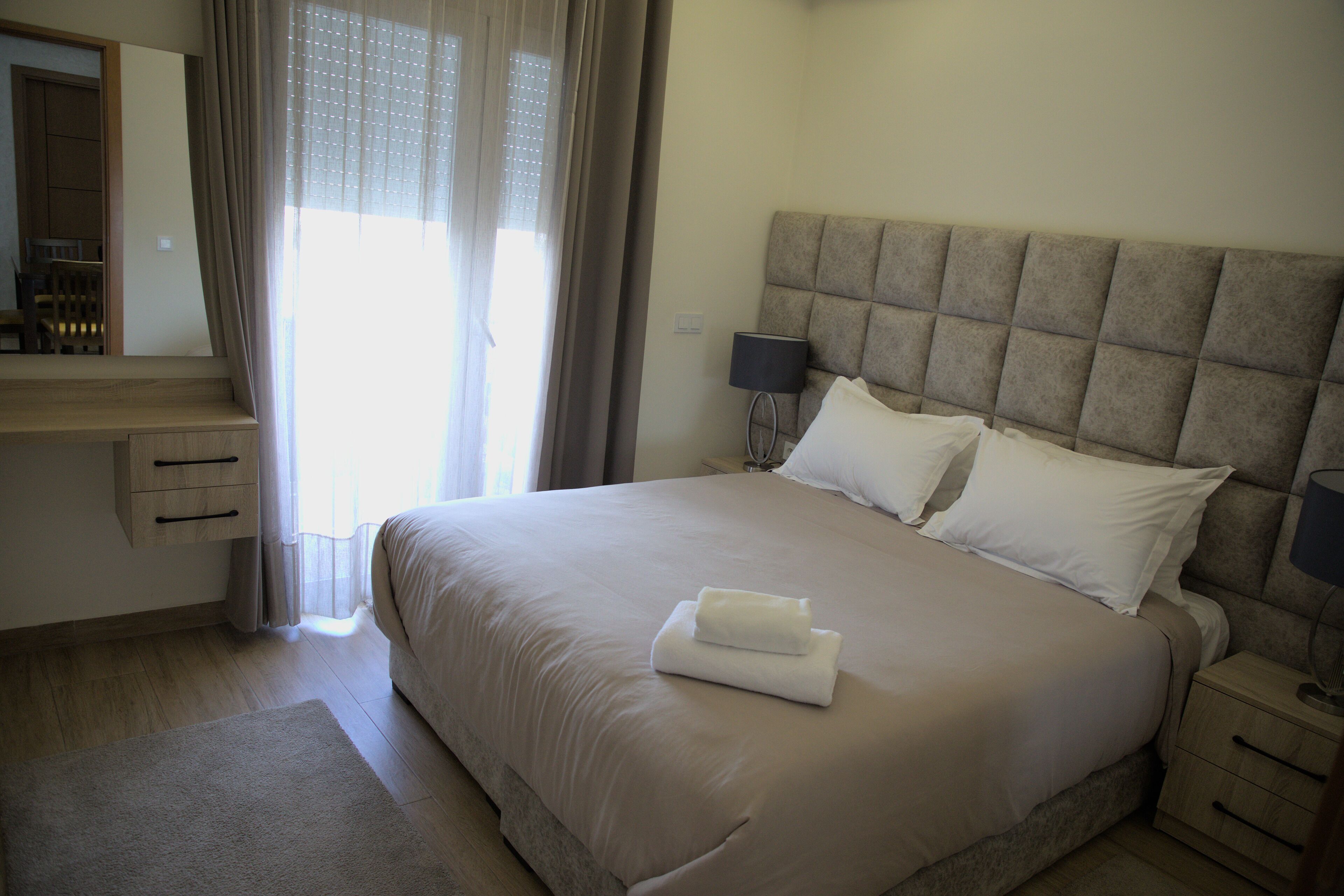 A neatly made bed with a headboard, a bedside table with a lamp, and a window with curtains.
