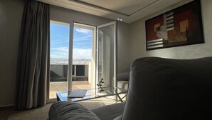 Un salon moderne avec un canapé gris, une table basse en verre et une vue sur le paysage urbain, visible à travers une porte-fenêtre coulissante.