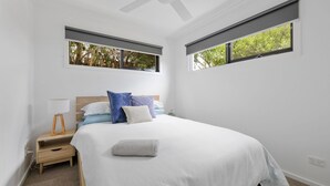 Un dormitorio con cama, mesita de noche, lámpara y dos ventanas con vistas al verdor.