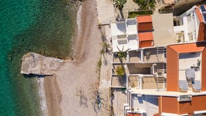 An aerial view of a coastal property with a beach, a rocky outcrop, and a multi-story building with red-tiled roofs.