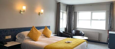 A hotel room with a bed, bedside table, lamp, chair, and a window with curtains.