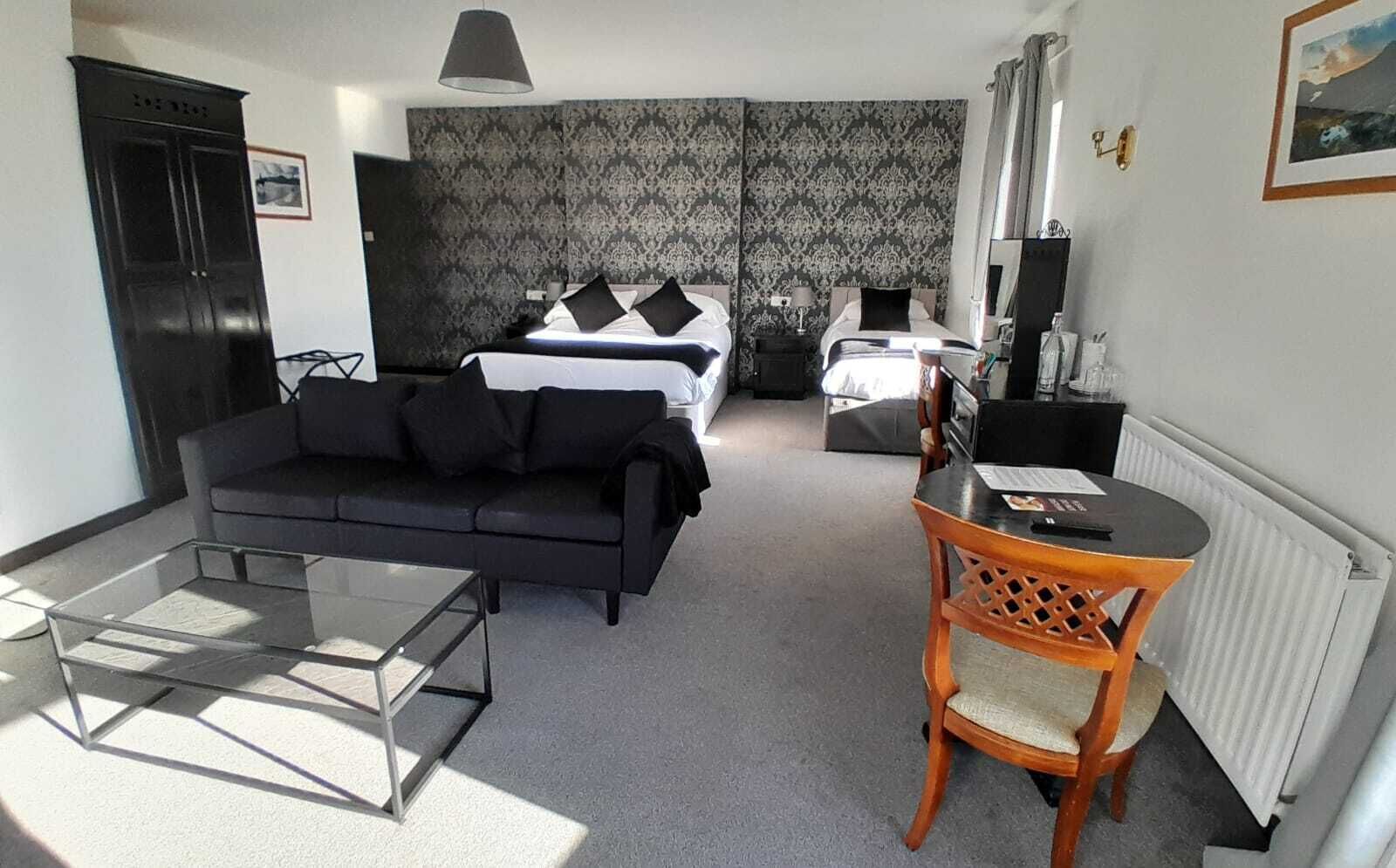 A modern hotel room with a black sofa, a small wooden table, a bed with black and white bedding, and a patterned wallpaper.