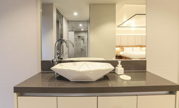 A modern bathroom with a large, geometric sink, a handheld soap dispenser, and a mirror reflecting a bed and a bathroom.