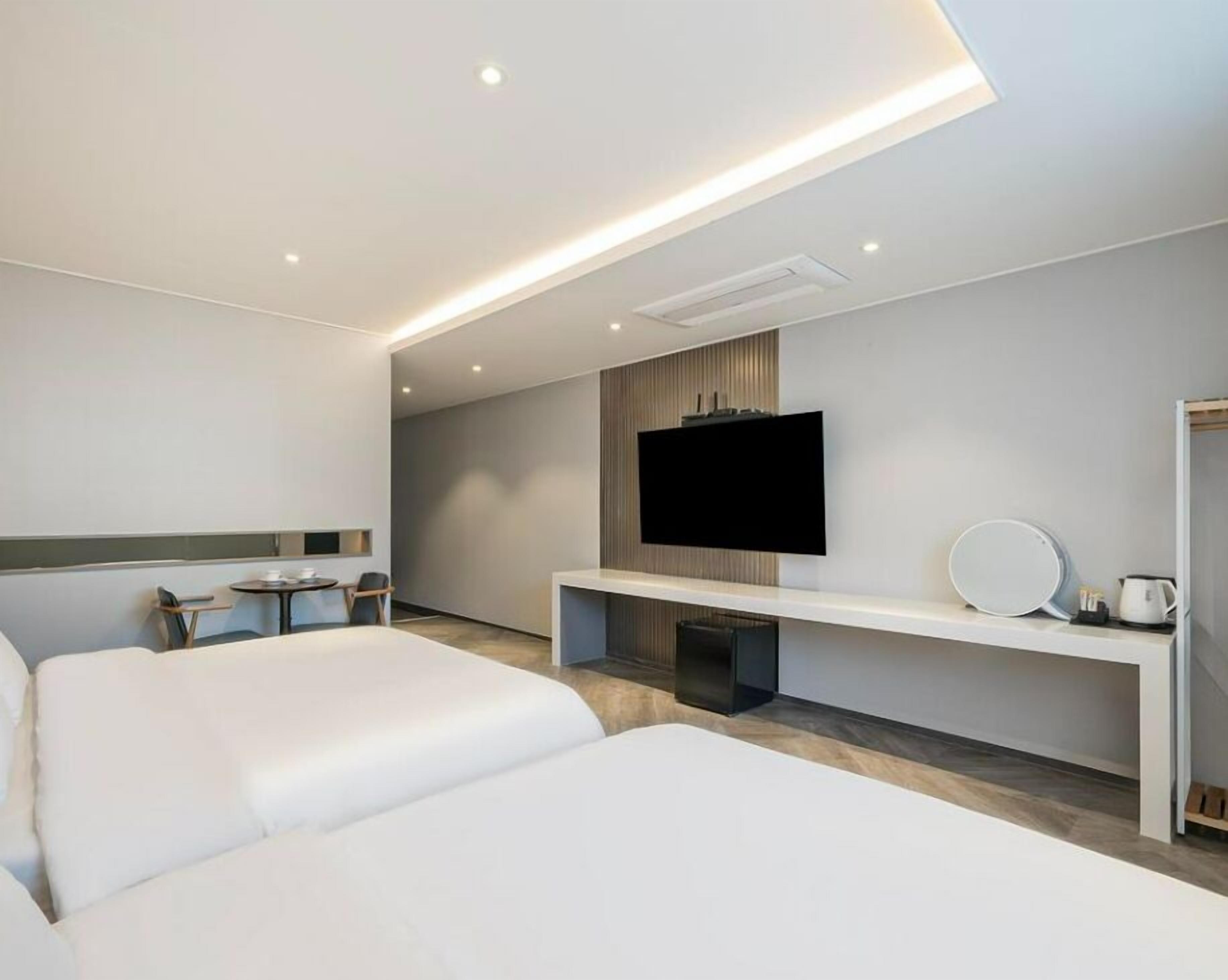 A modern hotel room with two beds, a flat-screen TV mounted on the wall, a small table with a chair, and a coffee machine.