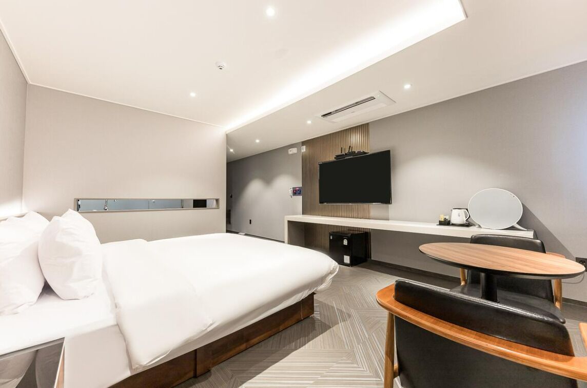 A modern hotel room with a large bed, a flat-screen TV mounted on the wall, a small round table, and a seating area with a chair.