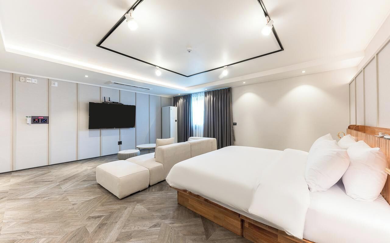 A modern hotel room with a large bed, a seating area, a flat-screen TV, and a control panel on the wall.