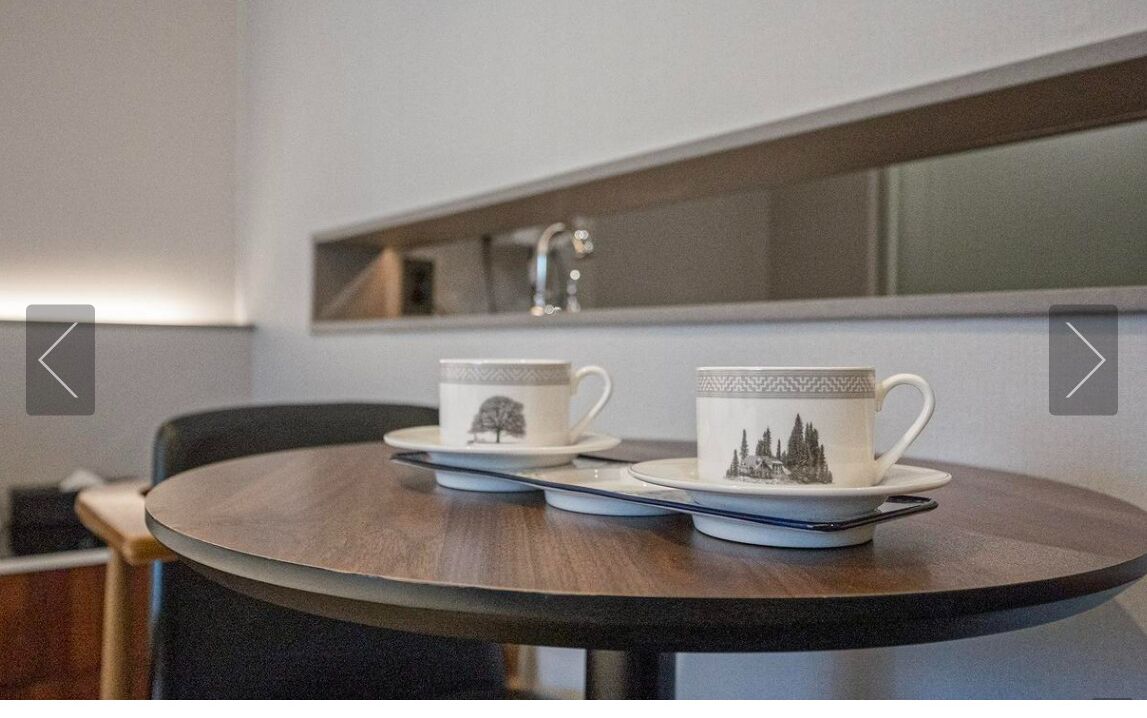 Ein runder Tisch mit zwei weißen Tassen mit schwarzen Mustern auf Untertassen, ein moderner Stuhl und ein an der Wand befestigtes Regal darüber.