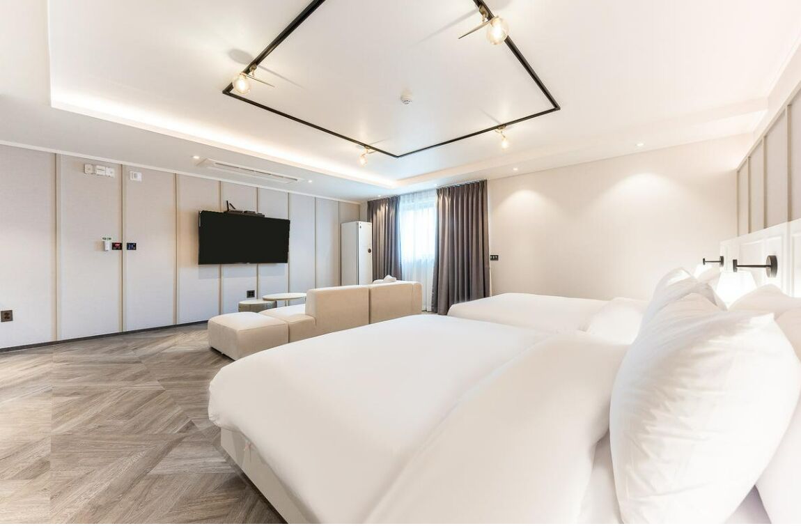 A modern hotel room with two large beds, a flat-screen TV, a sofa, and a wooden herringbone floor.