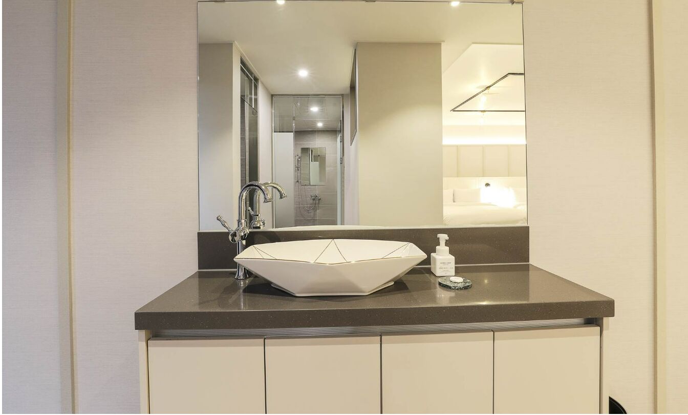 Een moderne badkamer met een grote spiegel, een geometrisch wit wastafeltje en een donker aanrecht.
