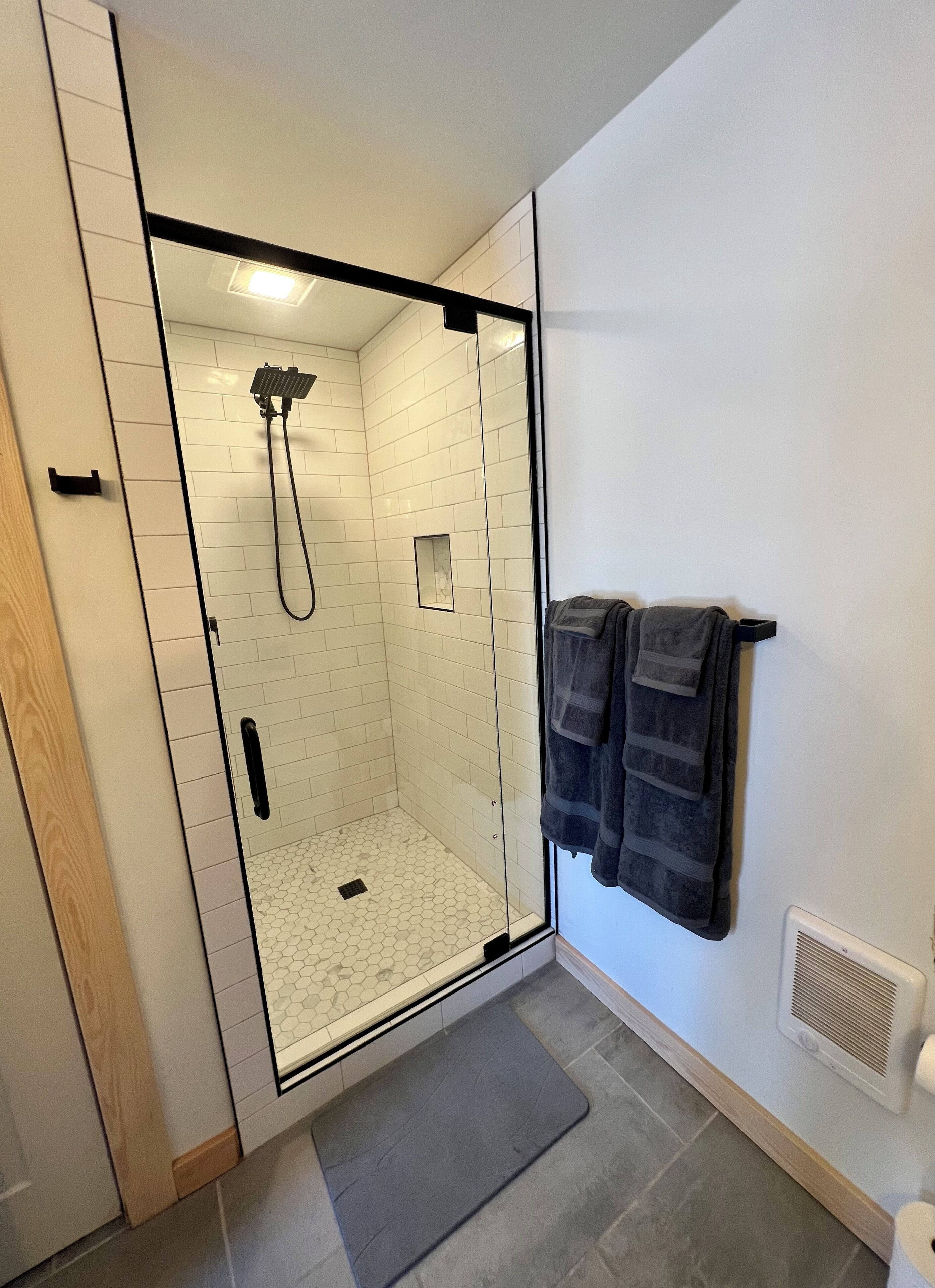 A walk-in shower with white tiled walls and a glass door, a towel rack with two towels, and a vent on the wall.