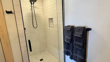 A walk-in shower with white tiled walls and a glass door, a towel rack with two towels, and a vent on the wall.