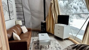 Un salon chaleureux avec un canapé en cuir marron, une table basse blanche, une commode blanche et un téléviseur. Une grande fenêtre offre une vue sur un paysage enneigé.