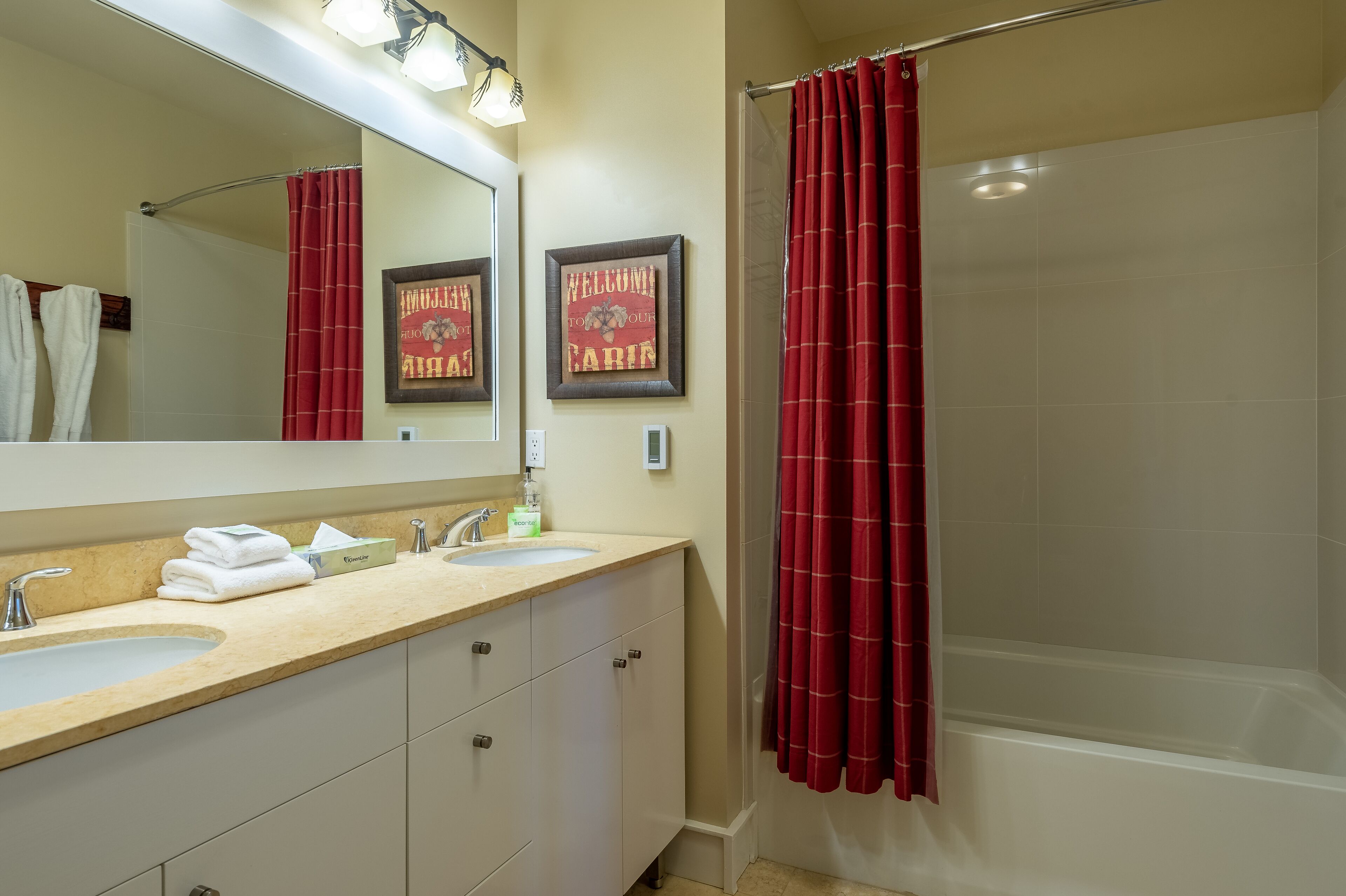 Une salle de bain avec deux lavabos, un grand miroir, une douche avec un rideau rouge et deux panneaux d’accueil encadrés.