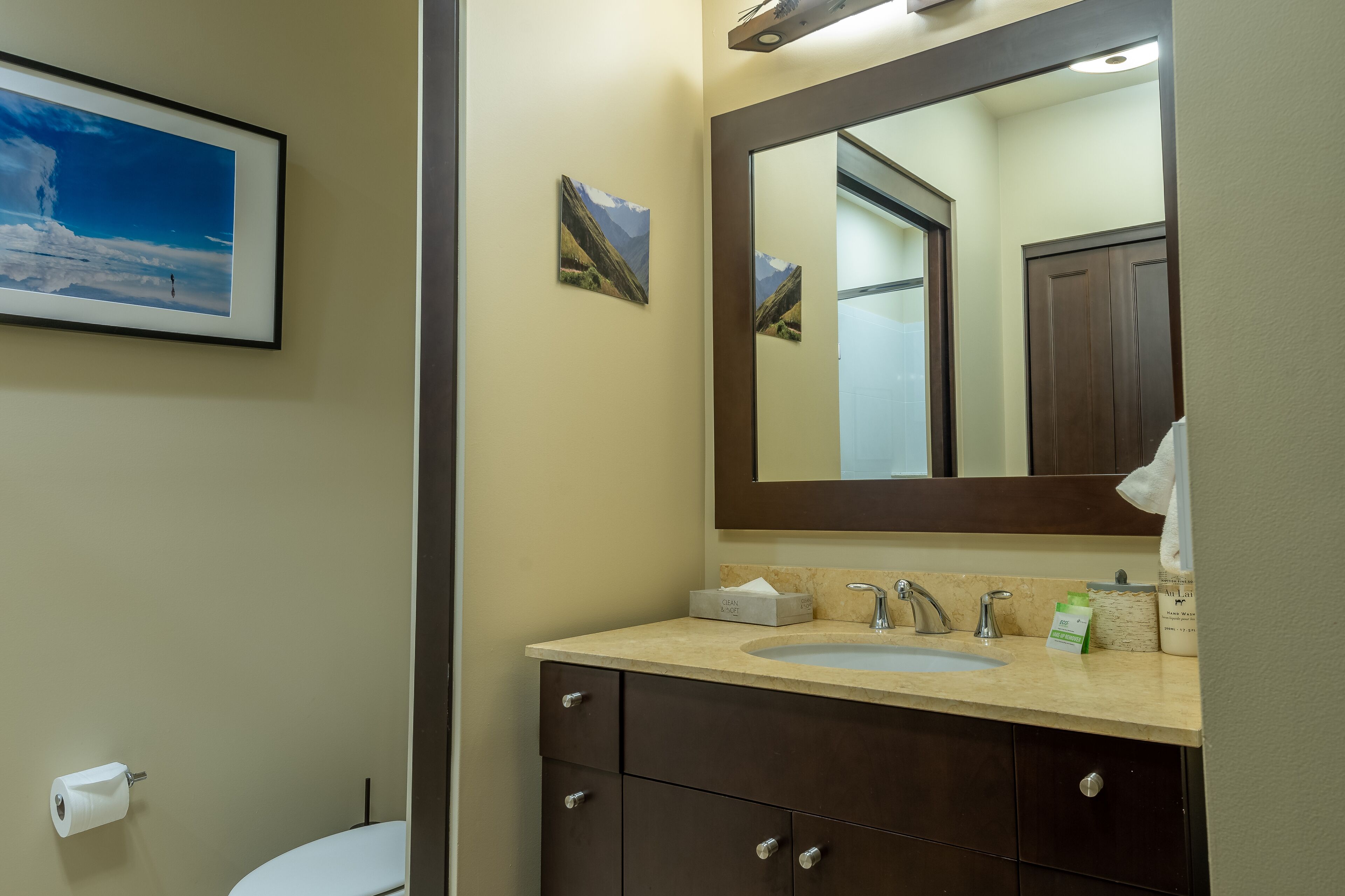 A bathroom with a sink, mirror, and toilet. There are framed pictures on the wall and a roll of toilet paper on the floor.