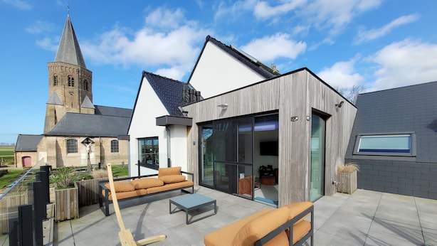 A modern outdoor patio with a sofa and coffee table, a wooden deck, and a view of a church tower. - b&b d'Ereplatse (Zuienkerke)