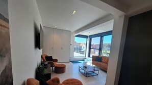 A modern living room with a sofa, ottoman, and coffee table. There is a large window providing a view of the outside. - b&b d'Ereplatse (Zuienkerke)