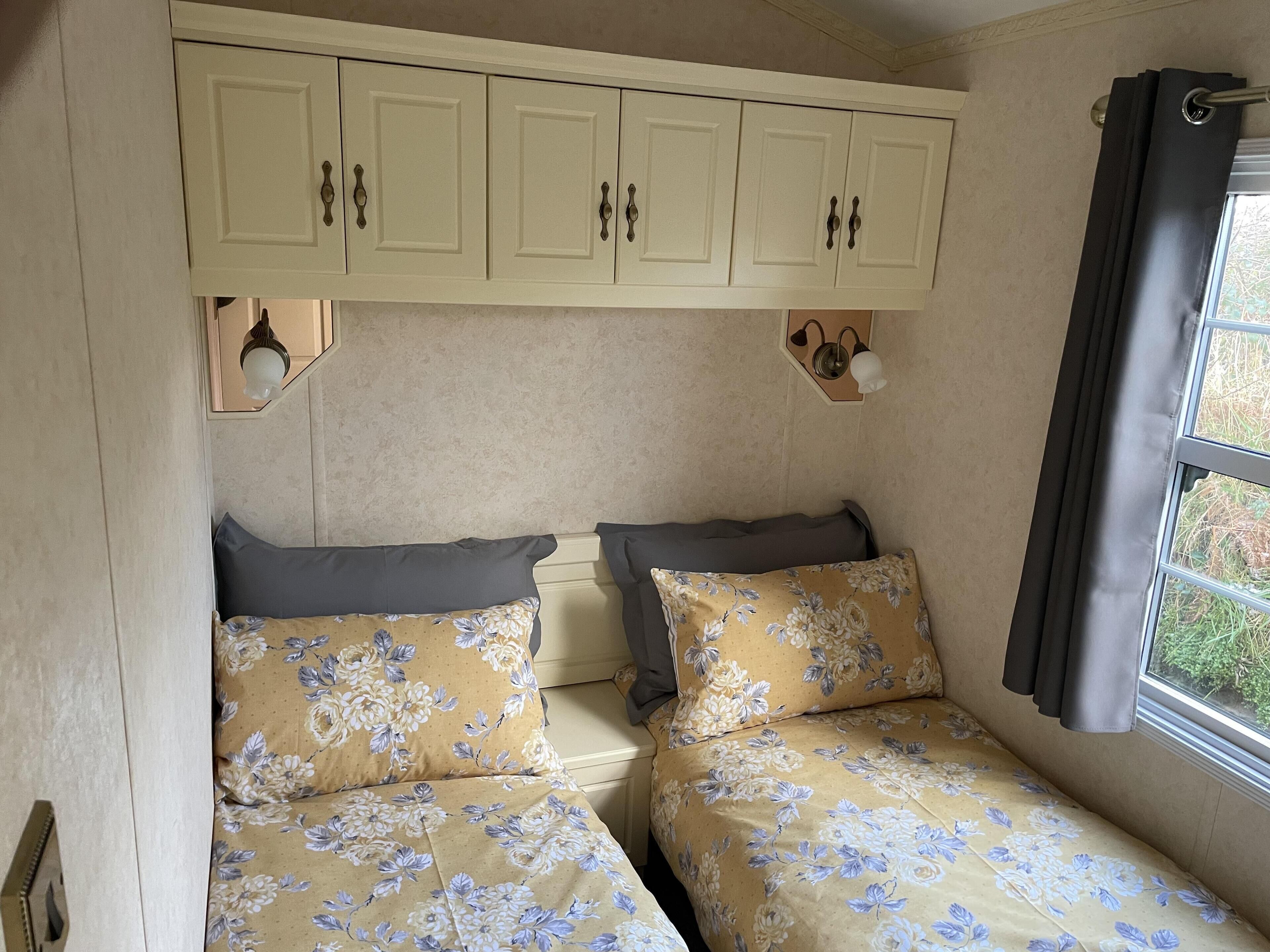 Une chambre de taille modeste, équipée de deux lits, chacun agrémenté de draps à motifs floraux, d’une petite fenêtre avec des rideaux et d’une armoire fixée au mur au-dessus des lits.