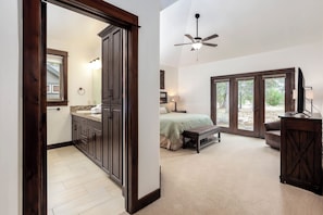 A bedroom with a bed, a nightstand, a bench, a ceiling fan, and a view through a large window. - TRAI39 - Stunning Caldera Springs Home (Sunriver)