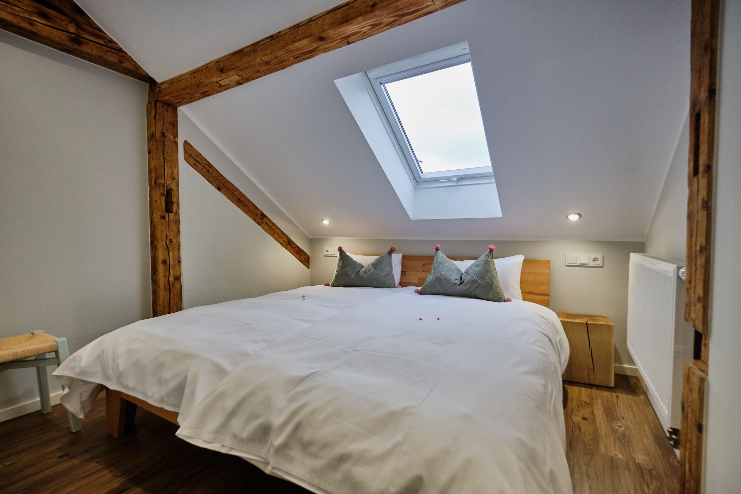 A bedroom with a skylight, wooden beams, a bed with pillows, a bedside table, and a radiator.