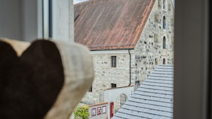 Vista a través de una ventana con un objeto en forma de corazón en primer plano, un edificio de piedra con tejado de baldosas al fondo y un letrero que indica un museo arqueológico.