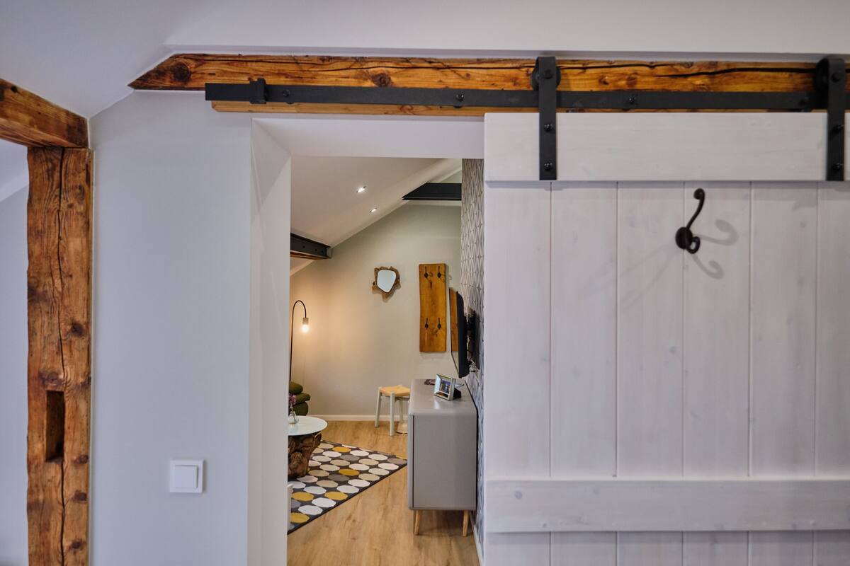 A room with a wooden ceiling beam, a white sliding door with a black handle, a small white table with a colorful rug, and a white cabinet with a mirror.