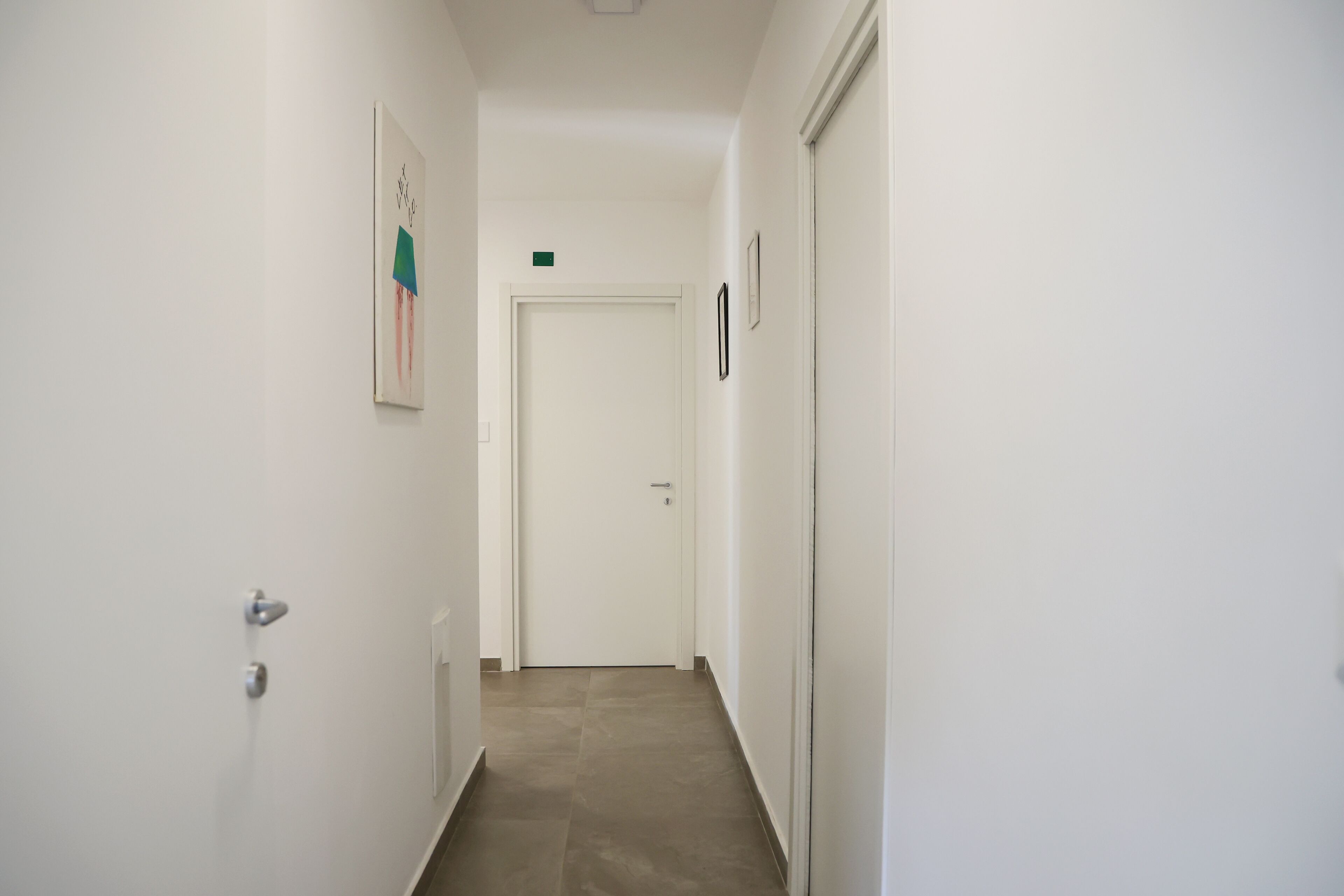 A narrow hallway with a white door at the end, a small green sign above the door, and a framed picture on the left wall.