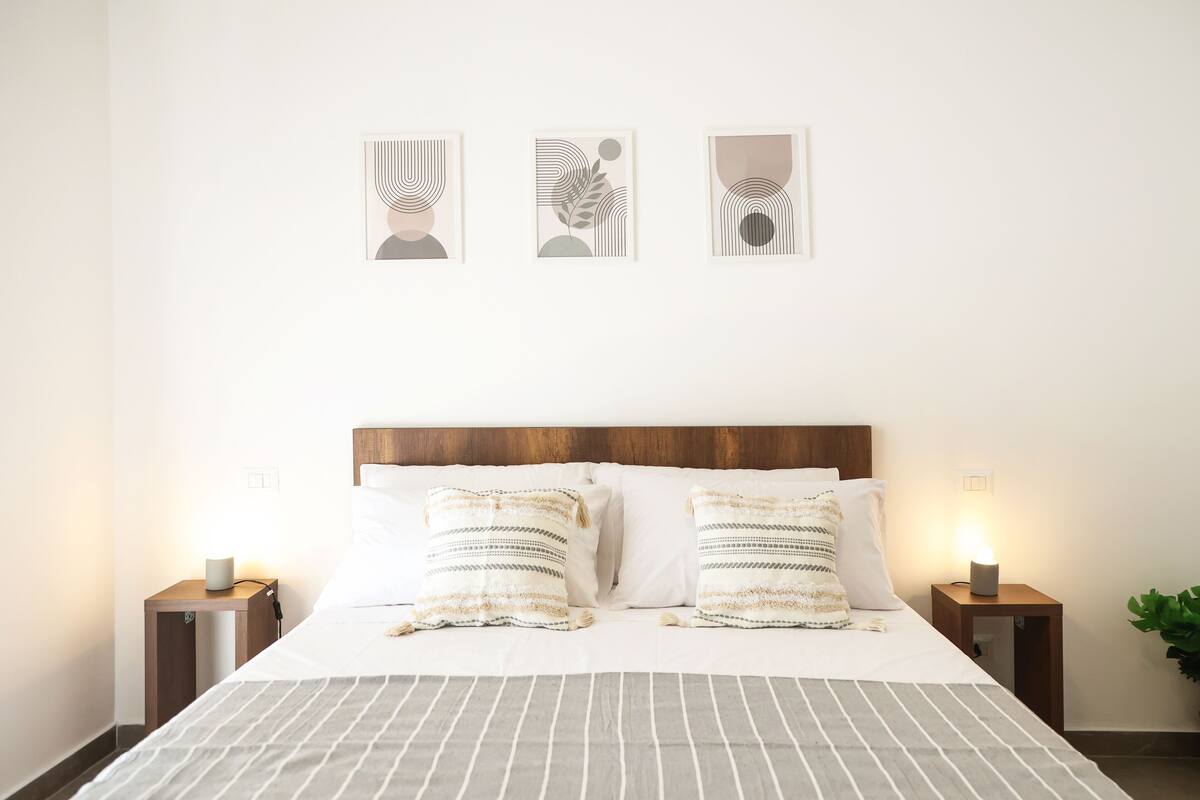 Una cama bien hecha con una colcha a rayas, dos almohadas y dos mesitas de noche con lámparas. Encima de la cama, hay tres obras de arte enmarcadas en la pared.