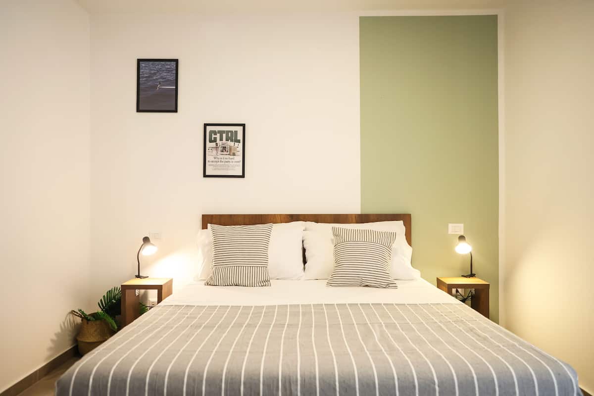 Un dormitorio ordenado con una cama, dos mesitas de noche con lámparas, un cuadro enmarcado y una pared verde decorativa.