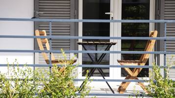 Un balcon avec une chaise en bois et une petite table, une rambarde en métal, et un bâtiment avec des volets et des fenêtres en arrière-plan.