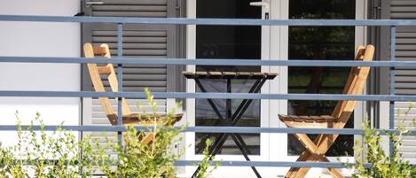 Een balkon met een houten stoel en een kleine tafel, een metalen hekwerk, en een gebouw met luiken en ramen op de achtergrond.