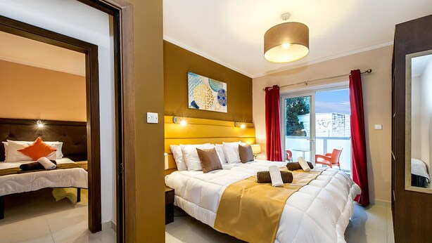 A hotel room with a large bed, a headboard, a painting, a window with curtains, and a balcony with chairs. - Blue 2: Two-Bedroom Apartment in St. Julian's (Swieqi)