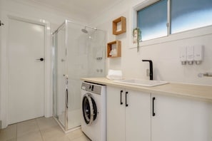 A modern kitchen with a glass shower enclosure, a built-in washing machine, and a sink with wooden shelves above.