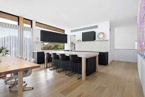 A modern kitchen with a central island, a dining area with a wooden table and chairs, and a large window with a view of greenery.