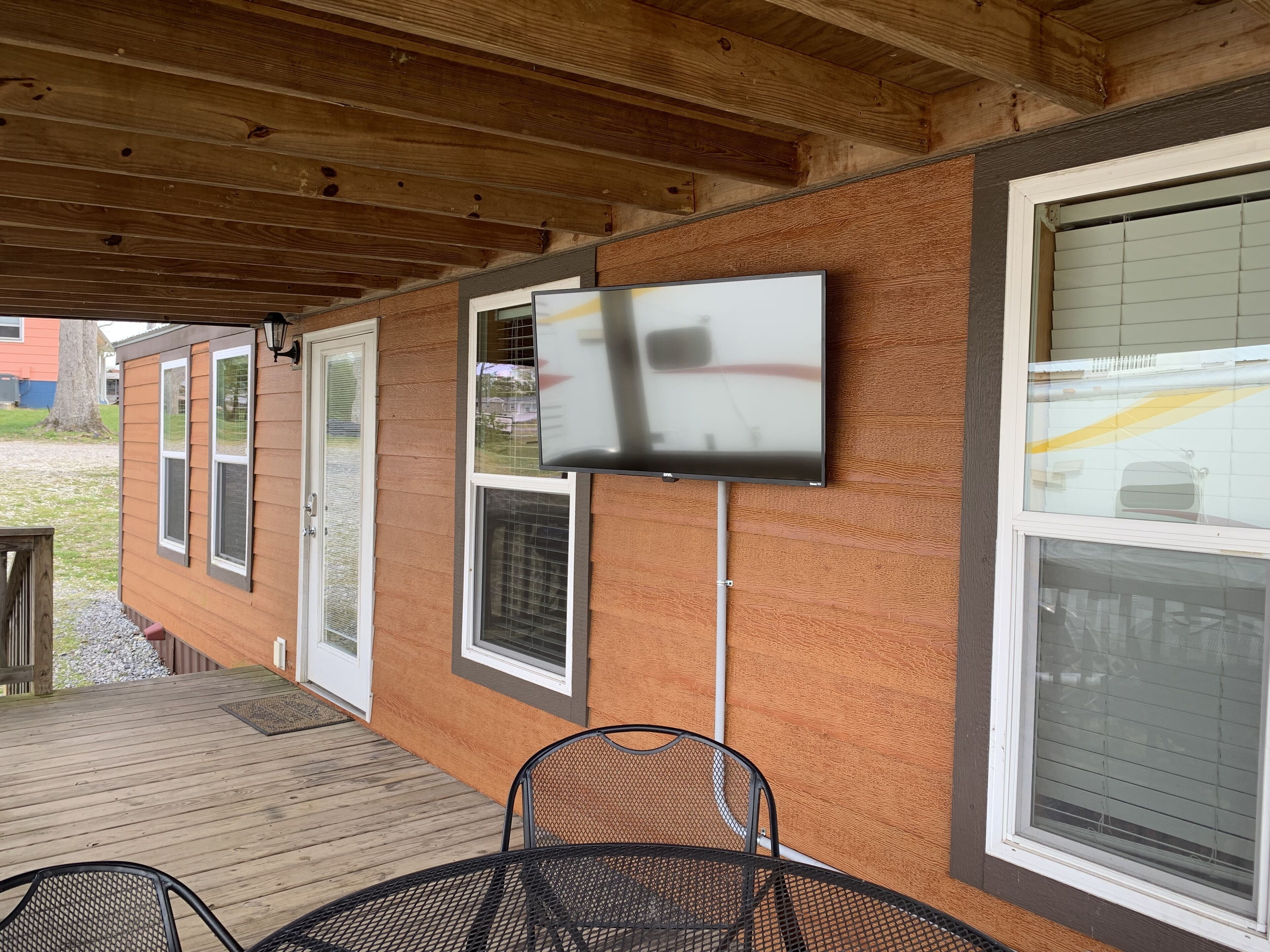 A covered porch with a table and chairs, a television mounted on the wall, and a window with blinds.