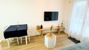 A modern hotel room with a wooden floor, a bed with pillows, a desk with a chair, a television on the wall, and a unique ceiling light fixture. - Magnifique F1 - à 50 de la mer (Bernières-sur-Mer)
