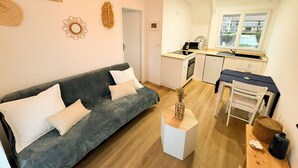 A compact living space with a grey sofa, a small white table, a kitchen area with a microwave, and a window with a view. - Magnifique F1 - à 50 de la mer (Bernières-sur-Mer)