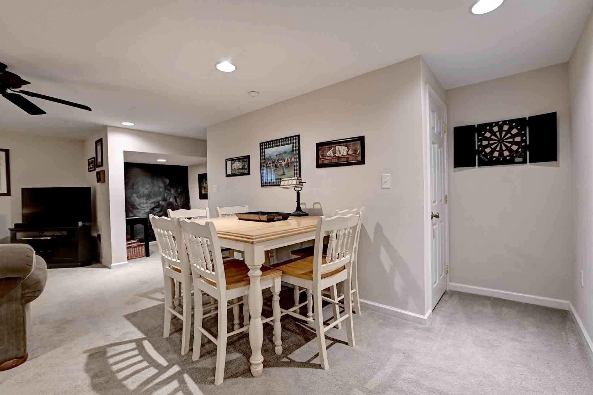 A dining area with a wooden table and white chairs, a ceiling fan, recessed lighting, and framed artwork on the walls.