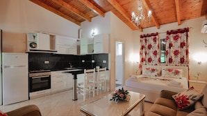 A modern kitchen with white cabinets, a black countertop, and a wooden ceiling. There is a dining area with a table and chairs, and a bedroom with a bed and floral curtains. - Villa Matilda (Lukovë)