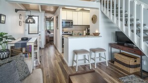 A modern kitchen with white cabinets, a central island with stools, and a staircase with white railings. - Beautiful Beachfront Bungalow (Lahaina)
